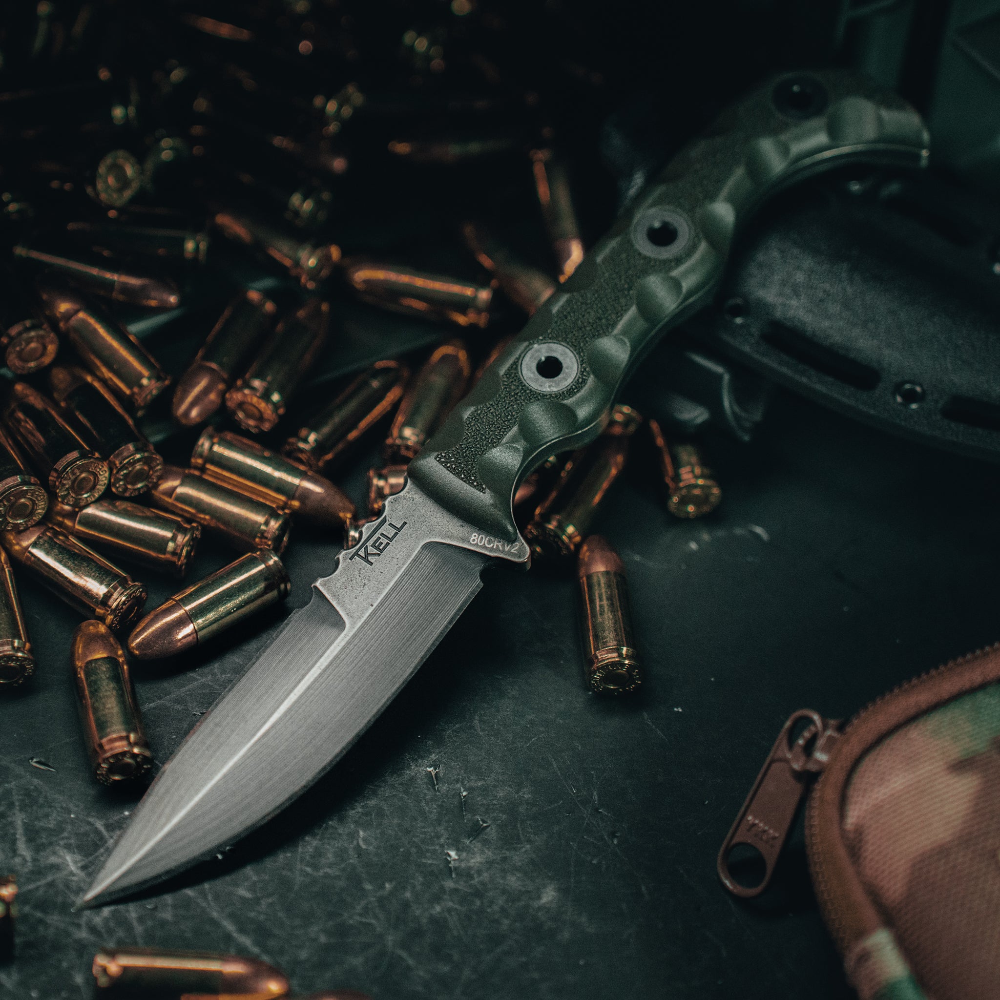 T.Kell AGENT 001 fixed blade tactical knife with od green battlelock grip resting among brass bullets on a dark surface